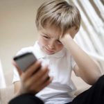 A photo of a young boy, who looks sad, sits looking at his smartphone in his hand whilst leaning his head in his other hand